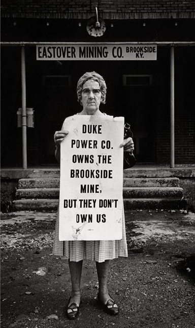 Woman with glasses wearing a dress in front of mining company holding sign.